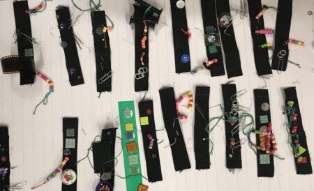 Several black fabric strips decorated with beads, buttons, film, pins, paper clips, and other small objects are arranged on lined paper.