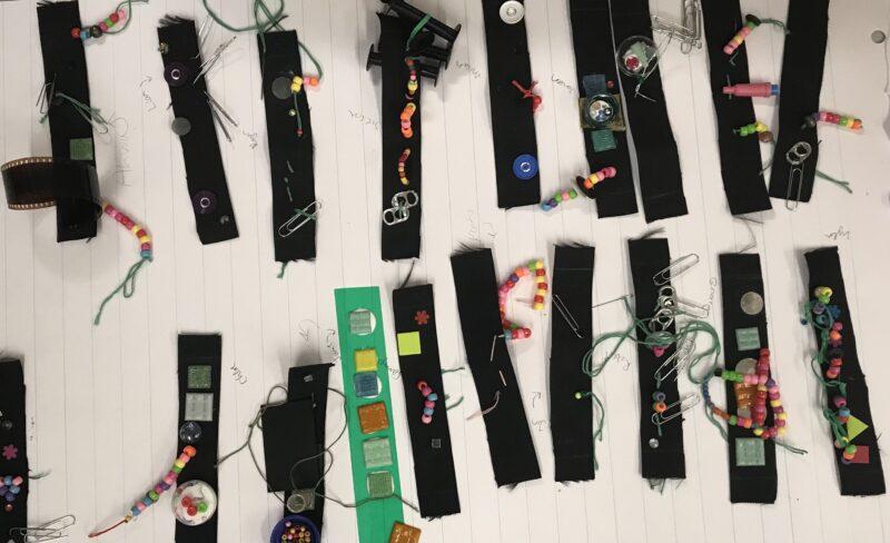 Several black fabric strips decorated with beads, buttons, film, pins, paper clips, and other small objects are arranged on lined paper.