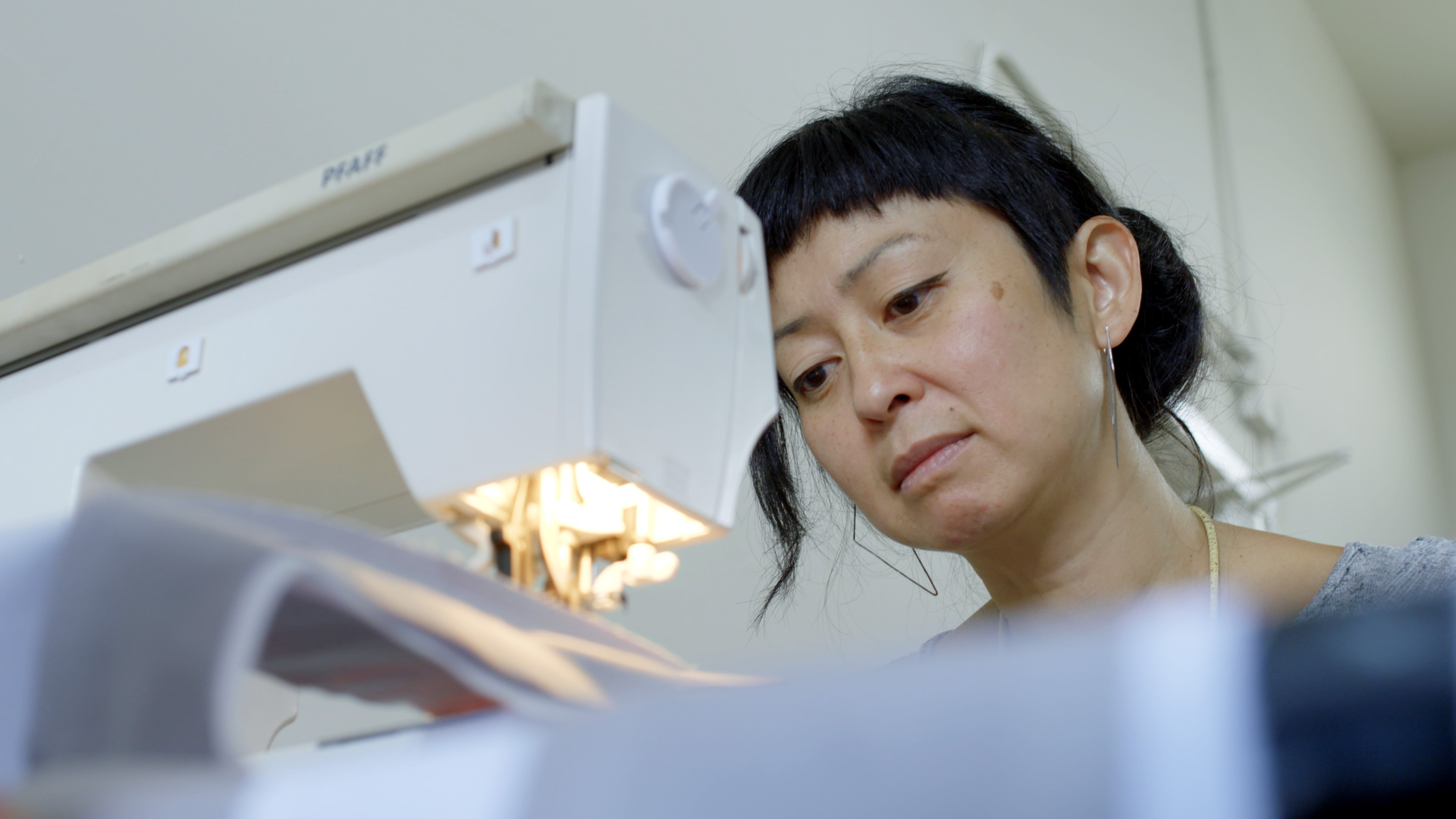 Stephanie Syjuco closely examines fabric as she works at a sewing machine in a brightly lit room.