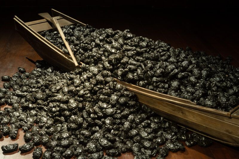 A sculpture that resembles a large pile of crabs spilling out of a small wooden fishing boat is installed on a dark wooden floor.