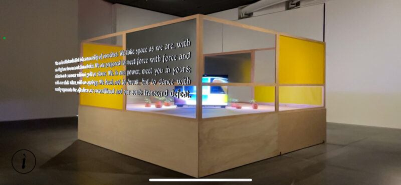A wooden-framed installation with yellow panels surrounds a TV and objects inside a gallery; white text is projected onto an outer panel.