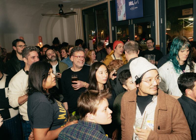 A large group of people stand closely together indoors, some holding drinks and looking toward something out of frame; the setting appears lively and social.
