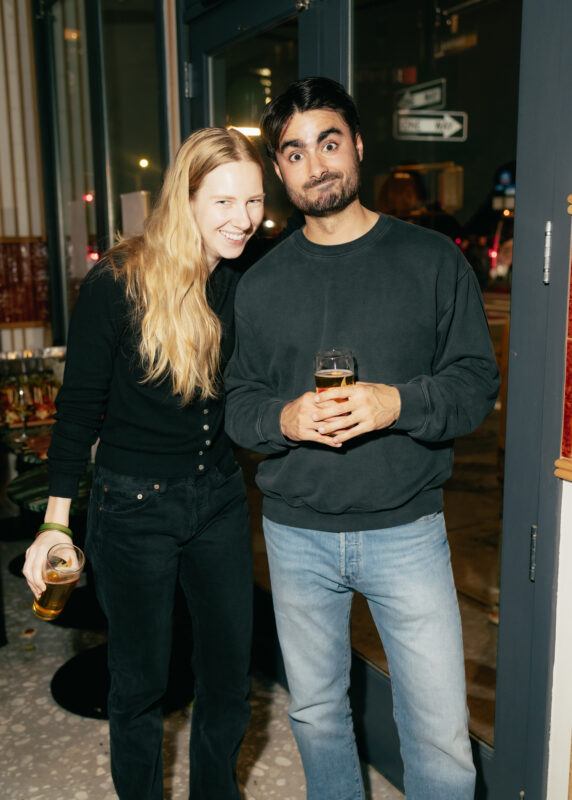 Two people standing indoors by a glass door, each holding a drink. The woman smiles at the camera while the man makes a playful face.