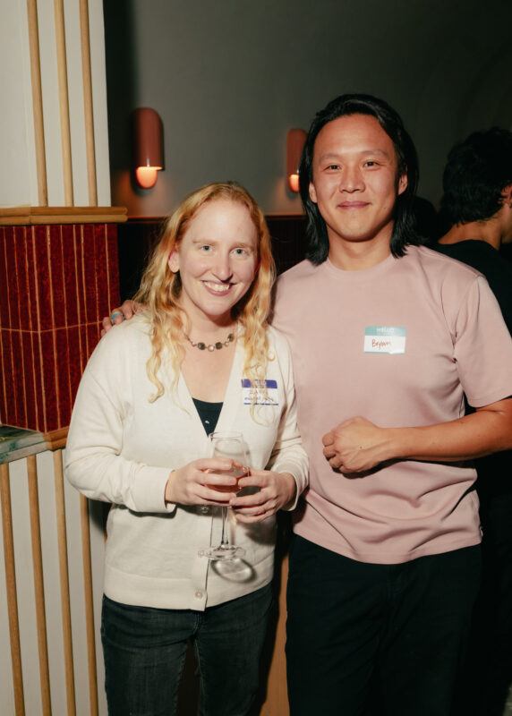 Two people stand indoors at an event; on the right a woman holds a wine glass, and both wear name tags and smile at the camera.