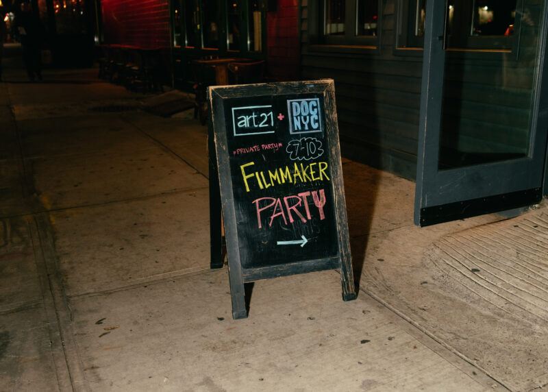 A chalkboard sign on a sidewalk reads "Filmmaker Party" with event details and an arrow pointing right.