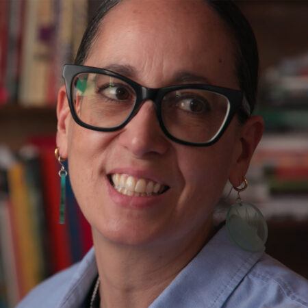 Dyani White Hawk wearing black glasses and earrings smiles while looking to the side, with a blurred bookshelf in the background.